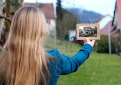 Frau hält einen Bilderrahmen mit einem Foto von einem Haus vor eine Baulücke